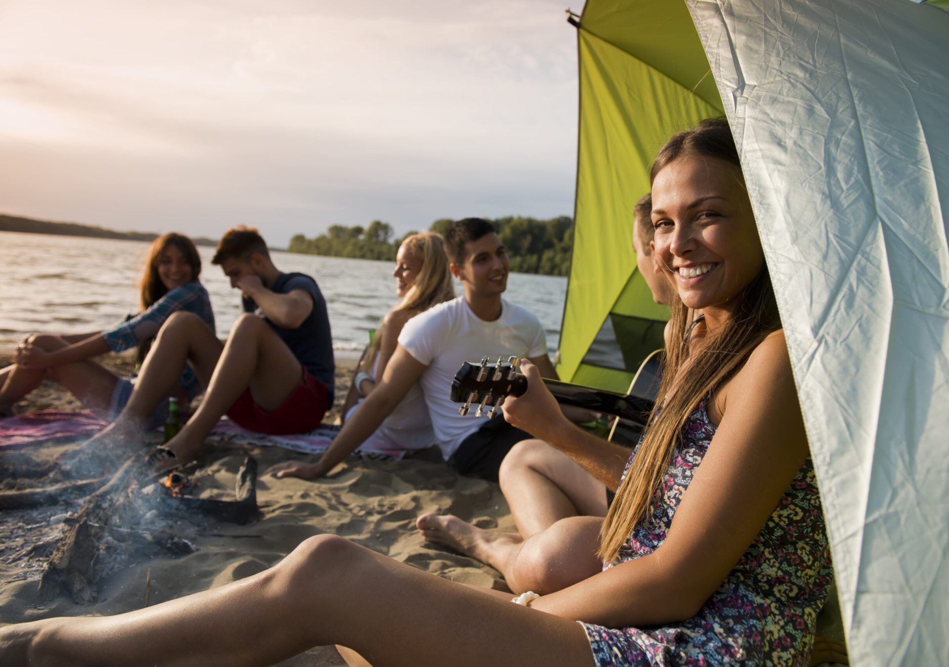 Friends enjoying in the sounds of the guitar, fire and sunset on the beach. Young woman looking at camera